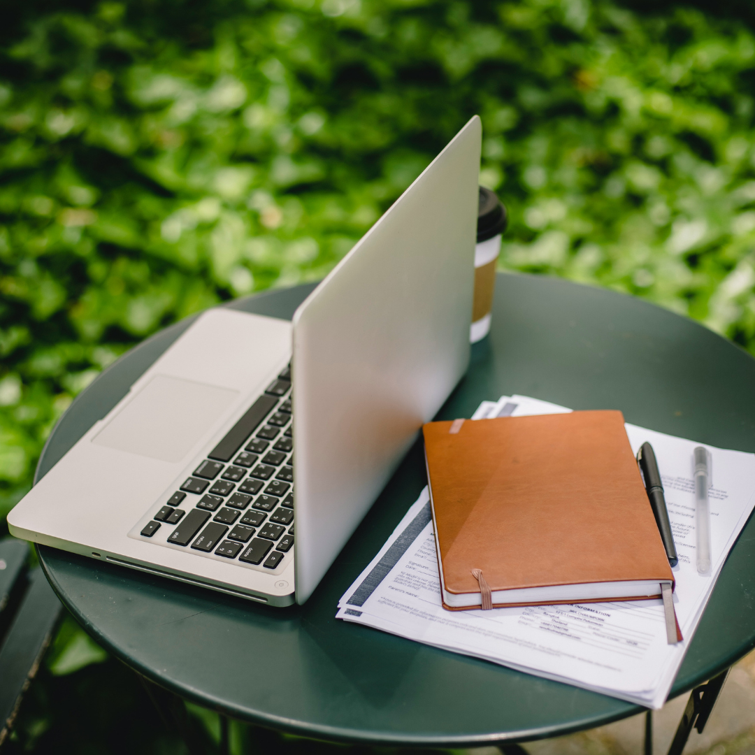Small outdoor table with a laptop, journal, pen, coffee, and papers, set outside.