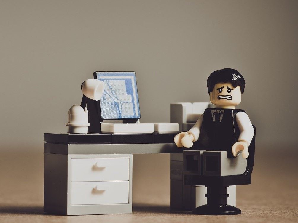 grimacing lego man sitting in business suite at a desk with computer, lamp