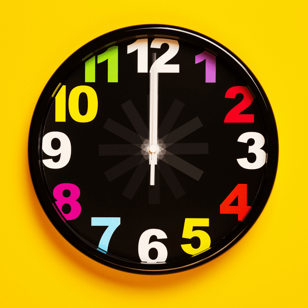 Black clock with multi color numbers and white minute and hour hands pointing to noon