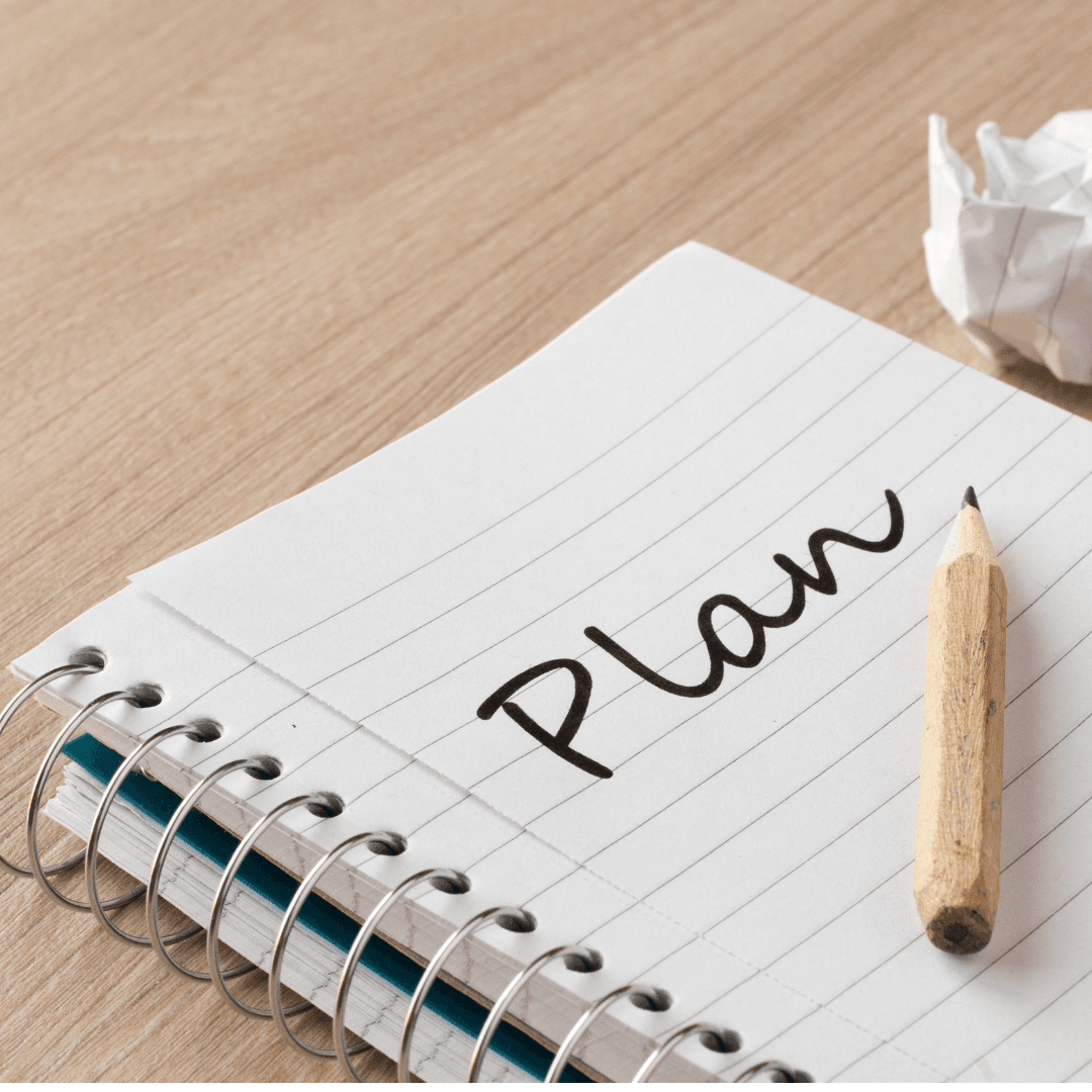 Open spiral notebook with the word Plan on a lined page and pencil on tabletop