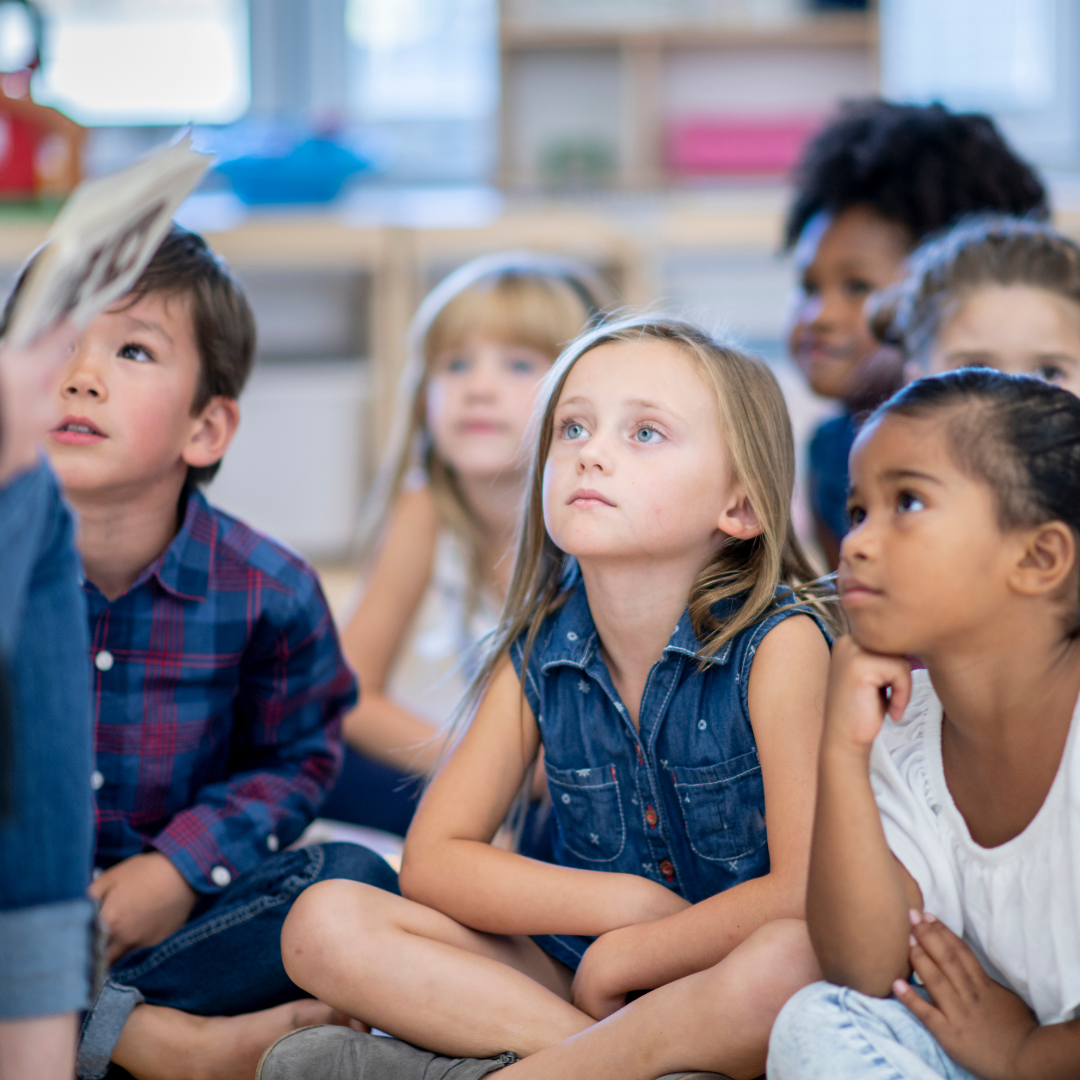 photo of children sitting on the floor cross legged, listening to someone read (not shown just leg)
